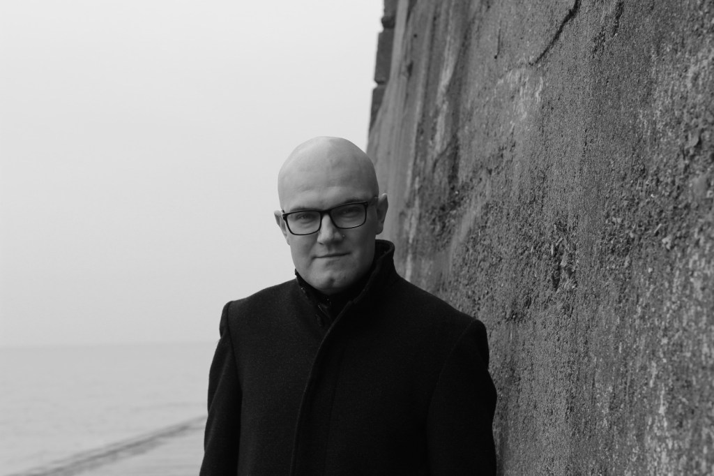 A black and white headshot of Martin Eve oudoors by the sea wearing a black coat and standing by a seawall. 