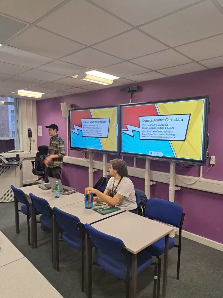 Two speakers prepare for the Comics Against Capitalism panel at the 22nd Annual Historical Materialism Conference in London. One person stands near the lectern speaking beside twin screens displaying the presentation title, while another sits at a table with notes and a drink can in front of a purple classroom wall.